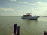 A classic white ferryboat is sailing on a calm, expansive body of water. The boat is adorned with string lights and a flag, and it has multiple decks filled with passengers. The sky is lightly cloudy, casting a soft glow over the scene. In the foreground, there are concrete posts near the water's edge.