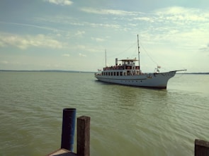 A classic white ferryboat is sailing on a calm, expansive body of water. The boat is adorned with string lights and a flag, and it has multiple decks filled with passengers. The sky is lightly cloudy, casting a soft glow over the scene. In the foreground, there are concrete posts near the water's edge.