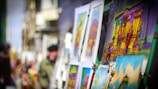 Several colorful paintings are displayed on easels in an outdoor setting. The paintings depict various scenes, including buildings and people in vibrant colors. A person with blurred features is in the background, possibly observing or arranging the artwork. The scene suggests an art market or outdoor gallery.