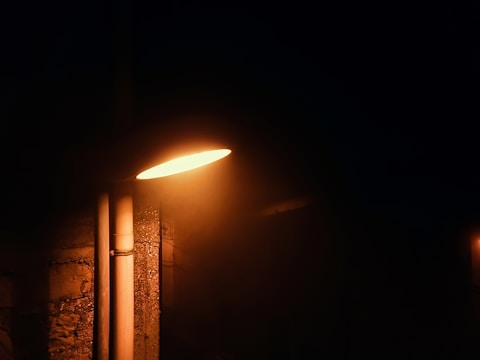 A streetlight illuminates a dark urban environment, casting a warm, orange glow. The surrounding area is dim, highlighting the textured surface of the wall and pipes next to the streetlight. Shadows and darkness dominate the background, offering a stark contrast to the light.