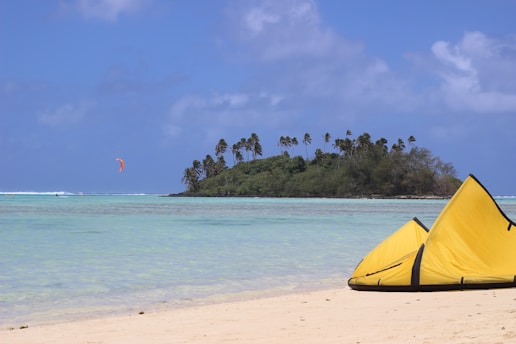 A tropical beach scene with a small island covered with palm trees in the background. The turquoise ocean is calm, and a parachute kite with a red color is visible in the distance. On the sandy beach in the foreground, two yellow kite surfing kites are laid down.
