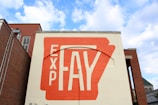 A large orange and white mural painted on the side of a brick building, with the text 'EXP FAY' prominently displayed. The mural covers the entire wall surface, surrounded by another red brick building on the left and a small green-roofed structure seen on the right. The sky is bright with scattered clouds.