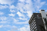 A modern multistory hotel building on the right with 'The Chancellor Hotel' signage. The sky is bright and filled with scattered fluffy clouds, creating a peaceful atmosphere. A few tree branches are visible in the foreground.