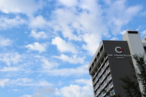 A modern multistory hotel building on the right with 'The Chancellor Hotel' signage. The sky is bright and filled with scattered fluffy clouds, creating a peaceful atmosphere. A few tree branches are visible in the foreground.