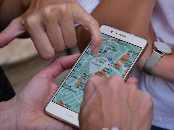 A smartphone displaying a digital map is being held by a hand, while another hand points to a specific location on the screen. Additional hands are partially visible, suggesting collaborative navigation or decision-making.