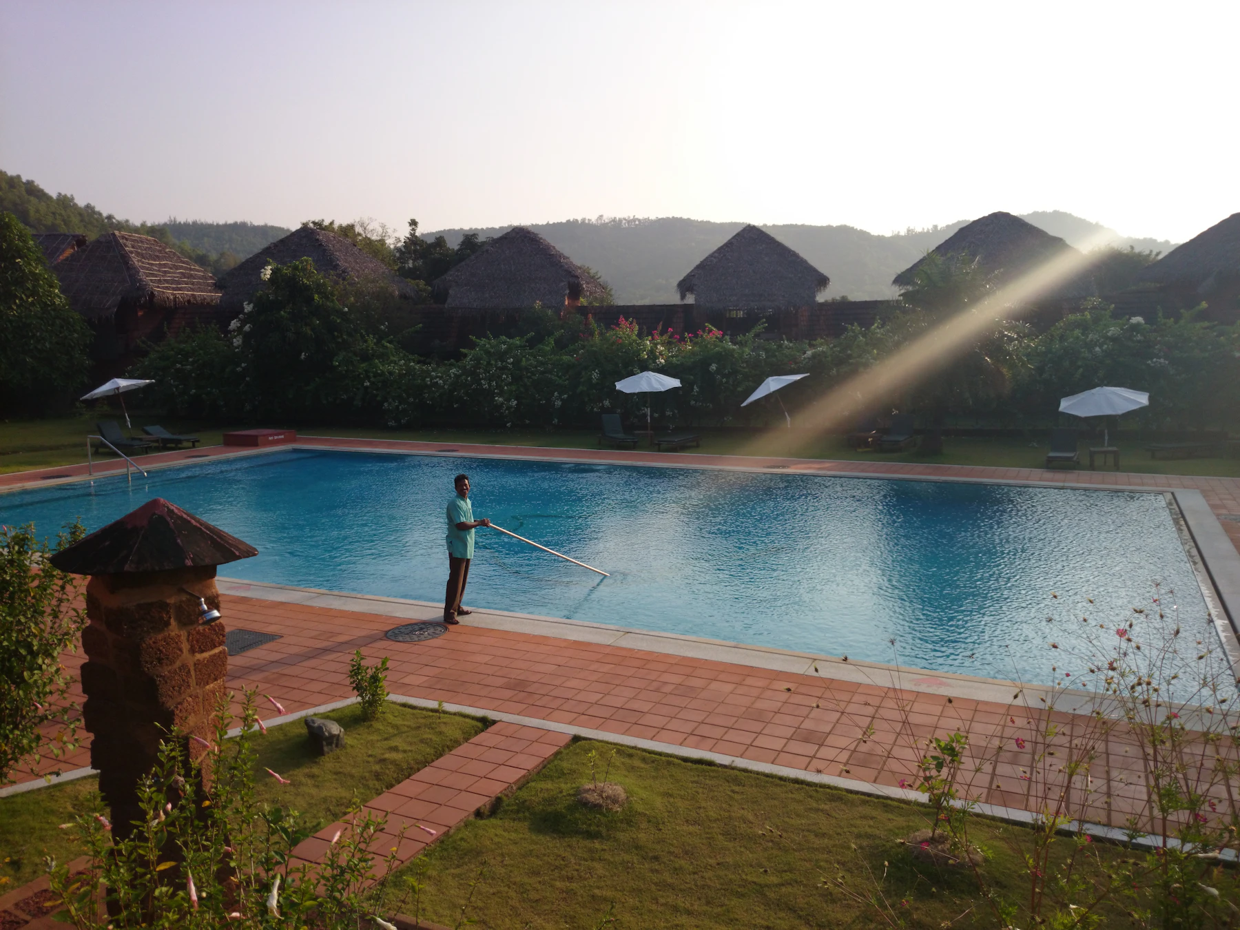 A man on a brick pool deck working a long pole across a large blue pool at sunrise