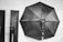 Black and white photography equipment setup featuring a large octagonal softbox and two rectangular softboxes against a brick wall. Lighting stands and wires are visible.