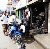 A group of men are gathered on a sidewalk in front of a small auto parts shop. A blue motorcycle is parked on the street beside them. The shop's open entrance reveals various mechanical parts and tools. Advertisements for auto parts are displayed on signs above the shops.