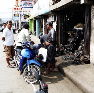 A group of men are gathered on a sidewalk in front of a small auto parts shop. A blue motorcycle is parked on the street beside them. The shop's open entrance reveals various mechanical parts and tools. Advertisements for auto parts are displayed on signs above the shops.