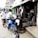 A group of men are gathered on a sidewalk in front of a small auto parts shop. A blue motorcycle is parked on the street beside them. The shop's open entrance reveals various mechanical parts and tools. Advertisements for auto parts are displayed on signs above the shops.