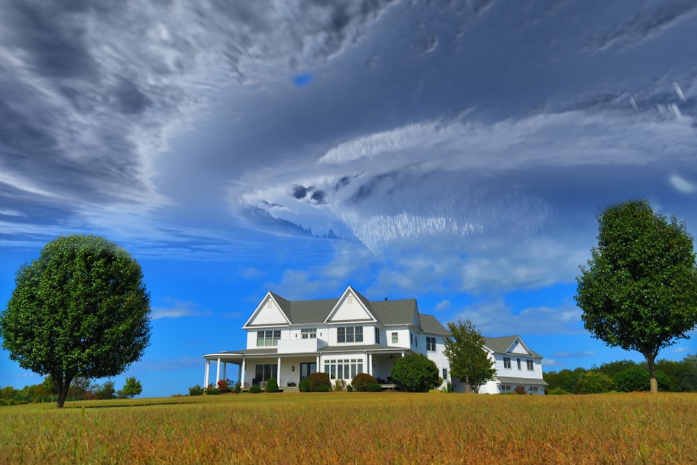 Maison blanche et grise entre deux grands arbres verts sous des nuages gris formant un tourbillon pendant la journée