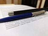 A close-up of two pens resting on top of printed text. The pens appear to be a sleek black pen with metallic accents and a blue pen with a silver tip. The text beneath them is printed on white paper, and the background suggests a desk or tabletop setting.