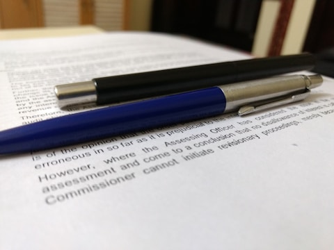 A close-up of two pens resting on top of printed text. The pens appear to be a sleek black pen with metallic accents and a blue pen with a silver tip. The text beneath them is printed on white paper, and the background suggests a desk or tabletop setting.