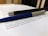 A close-up of two pens resting on top of printed text. The pens appear to be a sleek black pen with metallic accents and a blue pen with a silver tip. The text beneath them is printed on white paper, and the background suggests a desk or tabletop setting.