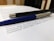 A close-up of two pens resting on top of printed text. The pens appear to be a sleek black pen with metallic accents and a blue pen with a silver tip. The text beneath them is printed on white paper, and the background suggests a desk or tabletop setting.