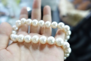 A hand holding a string of round, white pearls in a well-lit environment. The background is blurred, drawing focus to the pearls and the hand.