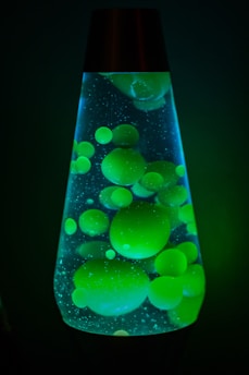 A lava lamp containing bright green blobs floating and moving inside a translucent liquid. The lamp is illuminated, creating a glowing effect in a dark setting, with bubbles and particles visible in the liquid.