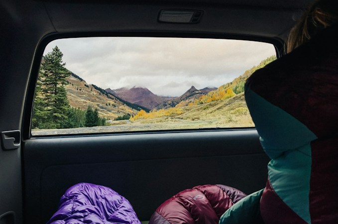 View from the back seat of a car through the rear side window to a mountain valley with autumn trees, purple and red sleeping bags piled on the seat below