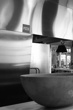 A black and white photo featuring a metallic pizza oven or kitchen equipment with a visible 'PIZZA' sign. In the foreground, there is a large round stone or ceramic bowl, and in the background, some shelving and a pendant light are seen in a modern interior.