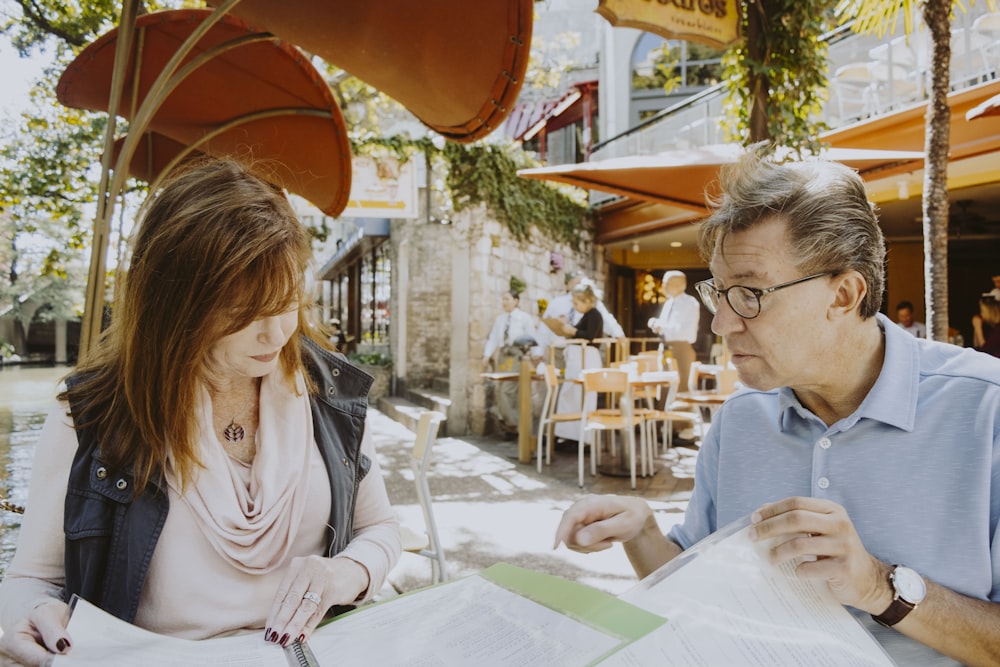 man and woman sitting down near table while reading outside cafe during daytime