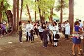 A group of people gather in an outdoor area surrounded by trees. Some are wearing lab coats, indicating a possible school or community event. Children and adults are engaged in conversation, and there is a general atmosphere of activity and interaction. In the background, a building labeled 'School Library' is visible.