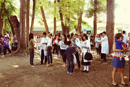 A group of people gather in an outdoor area surrounded by trees. Some are wearing lab coats, indicating a possible school or community event. Children and adults are engaged in conversation, and there is a general atmosphere of activity and interaction. In the background, a building labeled 'School Library' is visible.