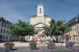A church with a clock tower surrounded by trees and colorful flowers in a quaint town square. The architecture is traditional with a clear blue sky in the background. A banner advertising a 'Tomato Festival' is visible in front of the church with additional signs. People are walking around the area, creating a serene and lively atmosphere.
