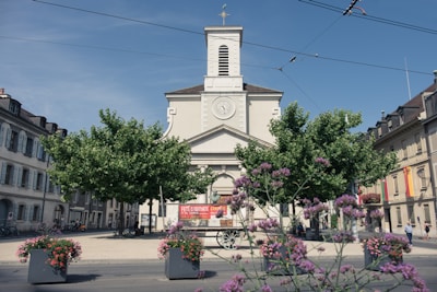 A church with a clock tower surrounded by trees and colorful flowers in a quaint town square. The architecture is traditional with a clear blue sky in the background. A banner advertising a 'Tomato Festival' is visible in front of the church with additional signs. People are walking around the area, creating a serene and lively atmosphere.