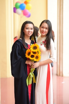 Two women pose closely together; one is wearing a graduation gown and holding a bouquet of sunflowers, while the other wears a white dress with red accents. In the background, colorful balloons are visible.