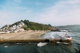 A coastal scene featuring a sandy beach, calm ocean waves, and a hilly area covered in greenery with numerous houses. In the foreground, there is a pier or seawall extending into the ocean with a red and white structure at the end. The shoreline curves gently with buildings and trees visible along the coast.