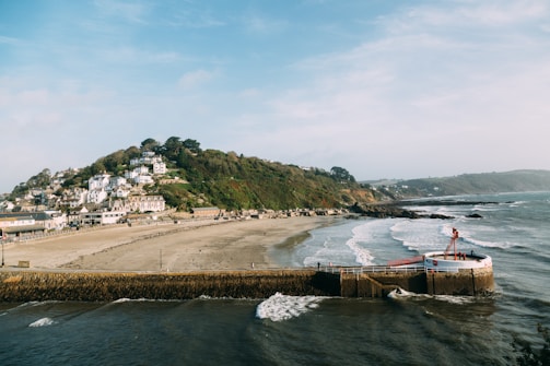A coastal scene featuring a sandy beach, calm ocean waves, and a hilly area covered in greenery with numerous houses. In the foreground, there is a pier or seawall extending into the ocean with a red and white structure at the end. The shoreline curves gently with buildings and trees visible along the coast.