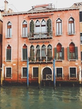 A historical Venetian building with Gothic-style windows and a pinkish-red facade, facing a canal. The architecture features ornate window arches and a small balcony decorated with potted plants. The building is reflected in the water of the canal below, which is a muted turquoise color.