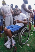 A man wearing sunglasses and a white t-shirt is sitting in a wheelchair, making a peace sign with his fingers. He is surrounded by a group of people, some standing and engaging with each other, while others are walking by. The setting appears to be outdoor with grass underfoot, suggesting a gathering or event.