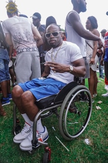 A man wearing sunglasses and a white t-shirt is sitting in a wheelchair, making a peace sign with his fingers. He is surrounded by a group of people, some standing and engaging with each other, while others are walking by. The setting appears to be outdoor with grass underfoot, suggesting a gathering or event.