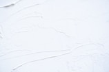 White textured surface with smooth and rough patches, possibly resembling thick paint or plaster applied onto a flat surface. The surface has visible brush or tool marks, creating a sense of movement and dimension.