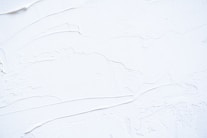 White textured surface with smooth and rough patches, possibly resembling thick paint or plaster applied onto a flat surface. The surface has visible brush or tool marks, creating a sense of movement and dimension.
