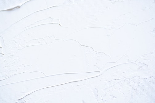 White textured surface with smooth and rough patches, possibly resembling thick paint or plaster applied onto a flat surface. The surface has visible brush or tool marks, creating a sense of movement and dimension.