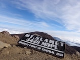 A rugged mountain landscape with a wooden sign displaying 'Mt. Slamet 3428 MDPL, Highest Point of Central Java'. The sky is bright and expansive with some clouds, while the foreground features rocky and dusty terrain.