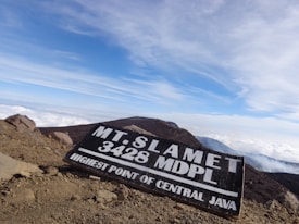 A rugged mountain landscape with a wooden sign displaying 'Mt. Slamet 3428 MDPL, Highest Point of Central Java'. The sky is bright and expansive with some clouds, while the foreground features rocky and dusty terrain.