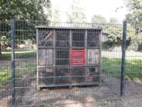 A large wooden structure resembling an insect hotel is enclosed within a metal fence. The structure is divided into several compartments filled with various natural materials like wood, twigs, and logs. It stands on a small area of soil surrounded by grass and trees in the background.