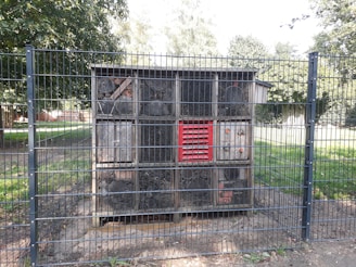 A large wooden structure resembling an insect hotel is enclosed within a metal fence. The structure is divided into several compartments filled with various natural materials like wood, twigs, and logs. It stands on a small area of soil surrounded by grass and trees in the background.