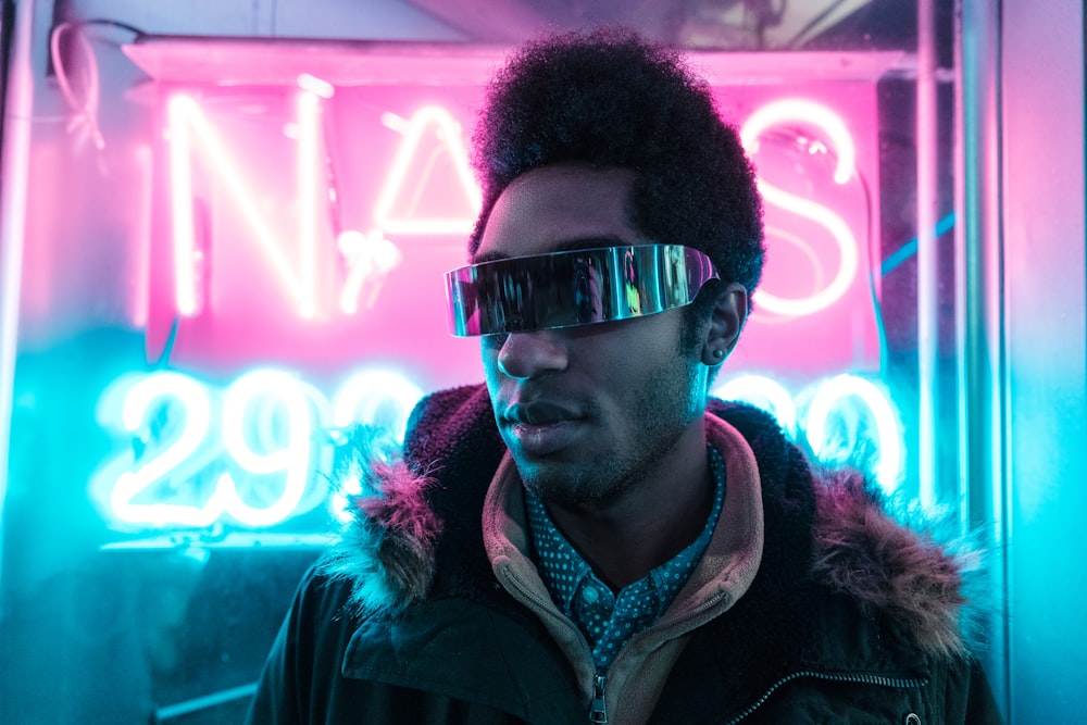 man wearing black parka jacket and black sunglasses in front of neon signage