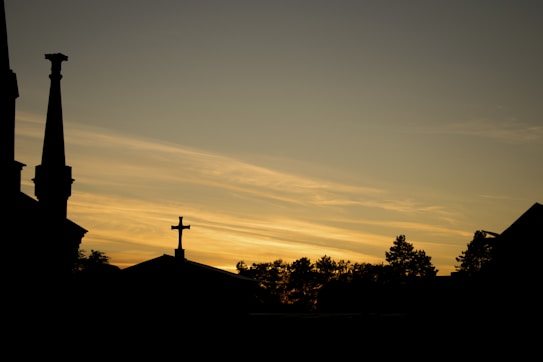 Silhouetted against a golden sky, church spires and a cross stand prominently. The background features a soft gradient of yellows and oranges, with scattered clouds reflecting the sunset. Trees form a darkened outline at the horizon.