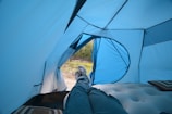 The interior of a blue tent with a view looking out towards a grassy landscape. A person is lying down on an inflatable mattress, with legs extended and feet visible, wearing blue pants and sneakers. There are striped pillows inside the tent.