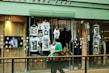 A shop window display featuring multiple white t-shirts printed with artistic black and white photographs. The storefront includes hanging t-shirts and several photographs attached to the glass. A television screen displays an image of a person in sunglasses. A person wearing a green shirt is walking past the display, holding a camera.