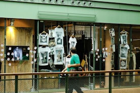 A shop window display featuring multiple white t-shirts printed with artistic black and white photographs. The storefront includes hanging t-shirts and several photographs attached to the glass. A television screen displays an image of a person in sunglasses. A person wearing a green shirt is walking past the display, holding a camera.
