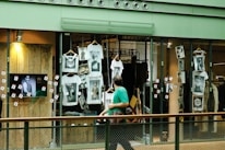 A shop window display featuring multiple white t-shirts printed with artistic black and white photographs. The storefront includes hanging t-shirts and several photographs attached to the glass. A television screen displays an image of a person in sunglasses. A person wearing a green shirt is walking past the display, holding a camera.