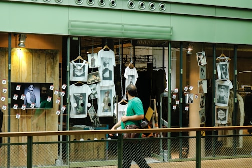 A shop window display featuring multiple white t-shirts printed with artistic black and white photographs. The storefront includes hanging t-shirts and several photographs attached to the glass. A television screen displays an image of a person in sunglasses. A person wearing a green shirt is walking past the display, holding a camera.