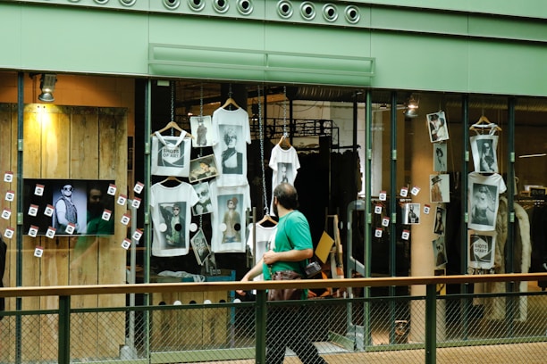 A shop window display featuring multiple white t-shirts printed with artistic black and white photographs. The storefront includes hanging t-shirts and several photographs attached to the glass. A television screen displays an image of a person in sunglasses. A person wearing a green shirt is walking past the display, holding a camera.