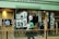 A shop window display featuring multiple white t-shirts printed with artistic black and white photographs. The storefront includes hanging t-shirts and several photographs attached to the glass. A television screen displays an image of a person in sunglasses. A person wearing a green shirt is walking past the display, holding a camera.